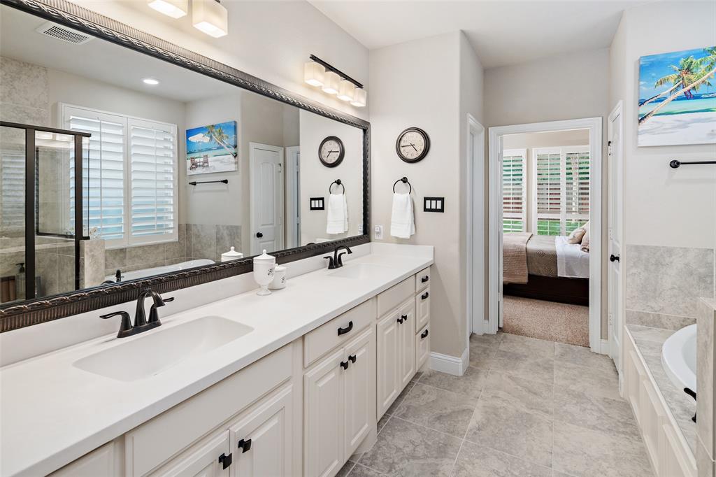 Dual-sink vanity with white countertops and matte black fixtures, full-width mirror with decorative trim, walk-in shower with glass enclosure, neutral tile flooring, and plantation shutters
