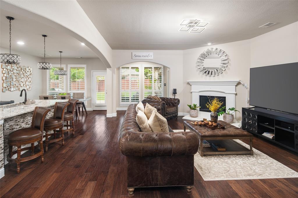 Open-concept living space featuring wood-finish flooring, a kitchen island with stone-finish countertops, a fireplace with a decorative mantel, and plantation shutters