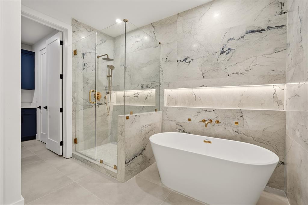 Freestanding oval soaking tub with metallic wall-mounted hardware, glass-enclosed walk-in shower with rainfall showerhead, floor-to-ceiling stone-patterned wall tiling, and integrated wall niches with accent lighting