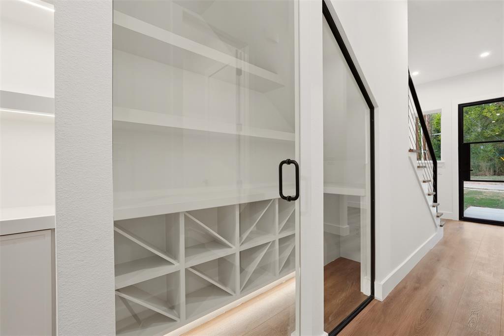 Built-in storage area featuring custom white shelving, integrated X-pattern wine rack, glass-paneled door with matte black hardware, recessed under-shelf lighting, and wood-finish flooring
