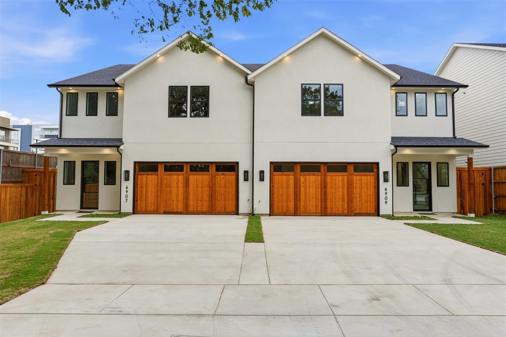 Duplex exterior featuring dual-pitched rooflines, smooth stucco siding, stained wood-finish garage doors with windows, and black-framed exterior lighting