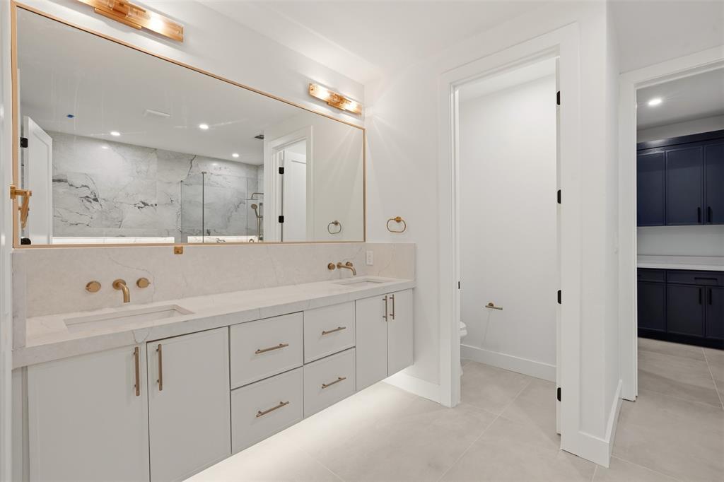 Floating vanity with stone countertop, dual undermount sinks, wall-mounted brass faucets, expansive framed mirror, and under-cabinet lighting