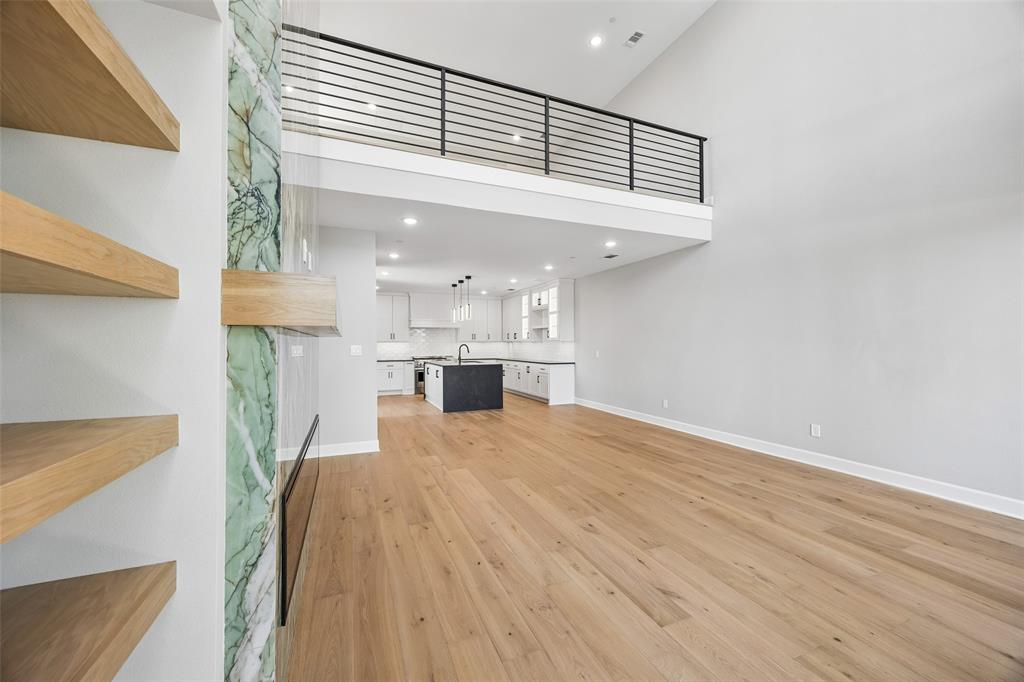 High-ceiling living area featuring wood-finish flooring, built-in wooden shelving, a floor-to-ceiling stone fireplace surround, and a lofted area with a metal railing