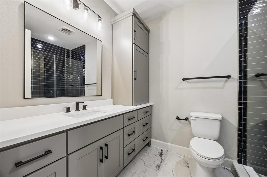Grey shaker-style cabinetry with matte black hardware, white countertop with integrated sink, rectangular mirror with thin dark frame, multi-light vanity fixture, and patterned tile flooring