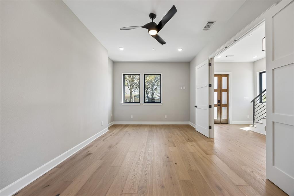 Light-filled interior space featuring wood-finish flooring, a modern black ceiling fan, dual windows with black frames, and white shaker-style double doors