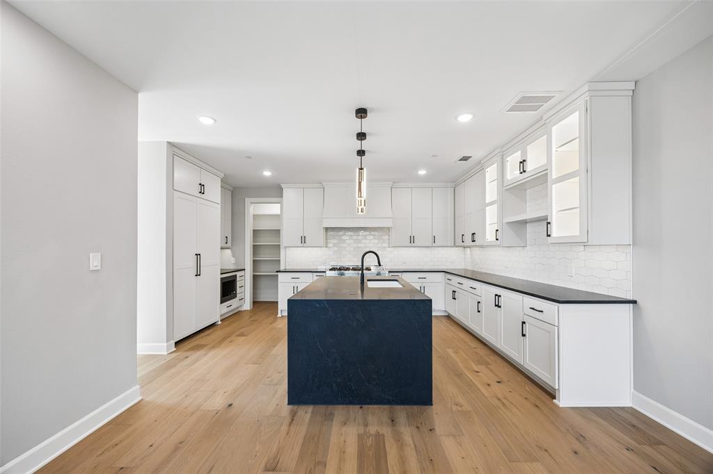 Kitchen featuring wood-finish flooring, a central island with a dark stone-surface countertop, white Shaker-style cabinetry, a tile backsplash, and built-in glass-front upper cabinets