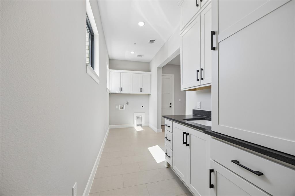 Laundry space featuring white shaker-style cabinetry, matte black hardware, integrated sink, dark solid-surface countertops, and large-format tile flooring