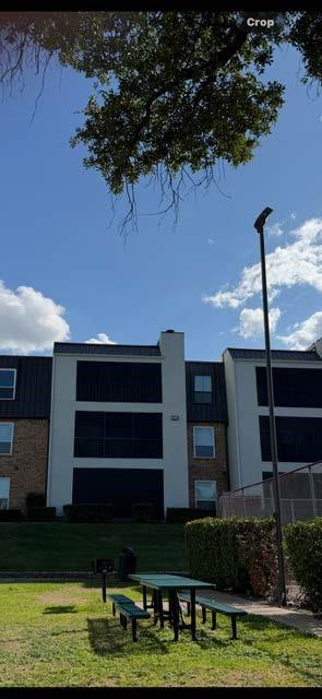 Multi-story residential structure featuring a contrasting white stucco and brick facade, dark metal roofing, and recessed balconies, situated on a grass-covered grounds area with a fixed outdoor grill and built-in bench seating