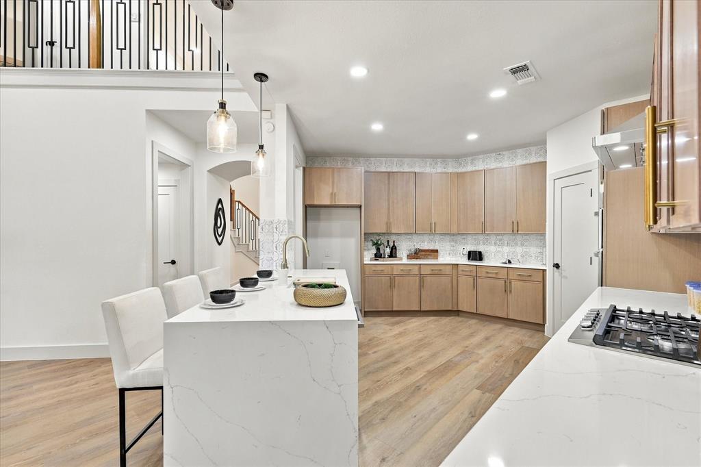 Kitchen featuring a waterfall-edge island with stone-surface countertop, flat-panel light wood cabinetry, decorative patterned tile backsplash, recessed lighting, and wood-finish flooring