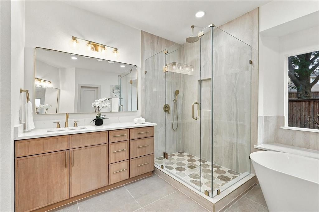 Wood-finish vanity with countertop, glass-enclosed shower featuring patterned tile flooring, wall-mounted faucet, and freestanding soaking tub