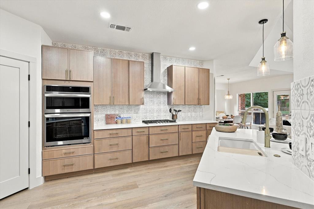 Kitchen featuring light-finish cabinetry, white countertops with veining, patterned tile backsplash, stainless steel built-in double oven and range hood, and wood-finish flooring