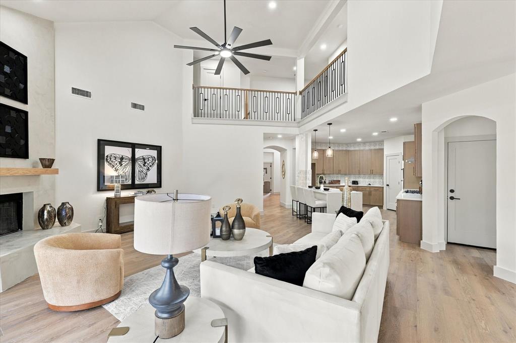 Double-height living area featuring a vaulted ceiling, wood-finish flooring, a fireplace with a light-tone hearth and wood mantel, a multi-blade ceiling fan, and a metal balcony railing
