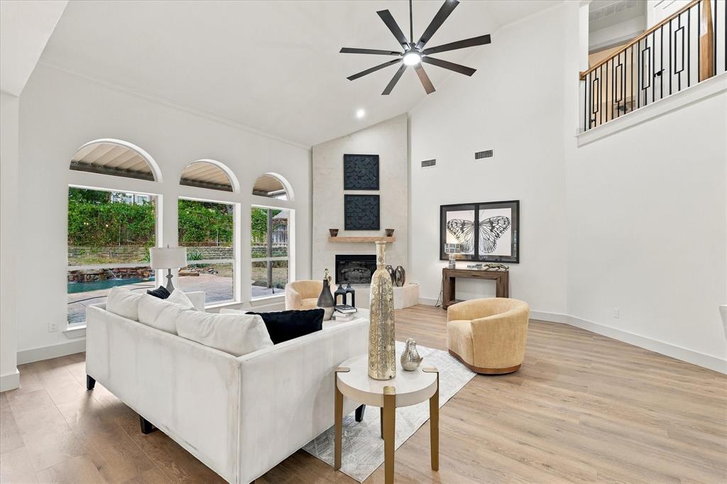 Vaulted ceilings with a multi-blade ceiling fan, expansive arched windows, wood-finish flooring, and a fireplace with a stone-surround facade featuring a wooden mantel