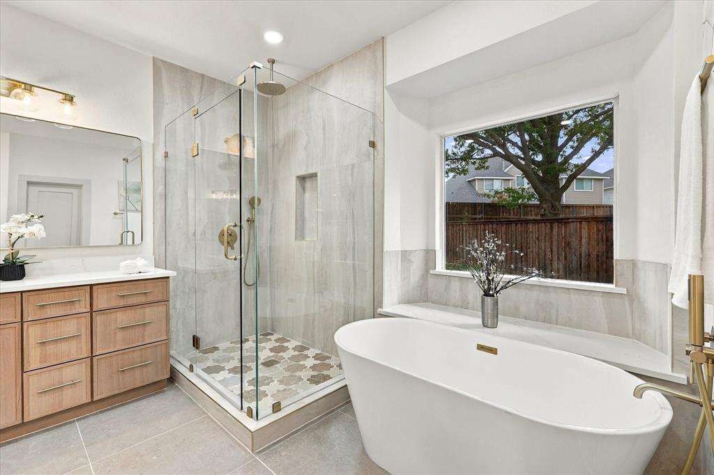 Freestanding soaking tub, glass-enclosed walk-in shower with stone tile surround, patterned tile shower floor, light wood-finish vanity, and picture window