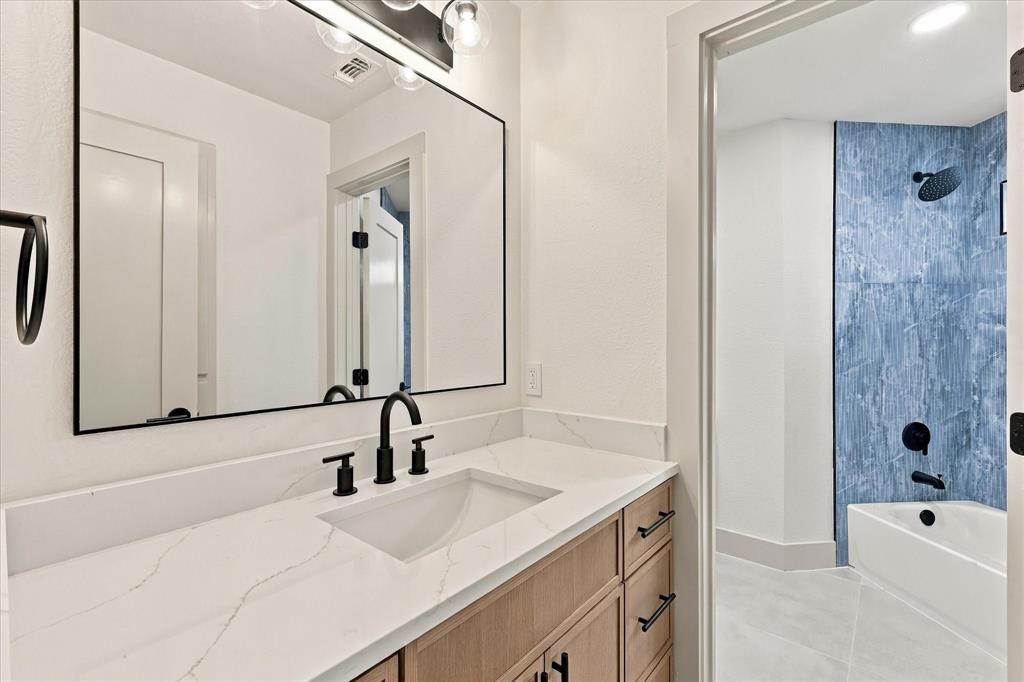 Bathroom featuring a vanity with a white stone-look countertop, wood-finish cabinetry, a large rectangular mirror, and matte black hardware including a wall-mounted light fixture and widespread faucet