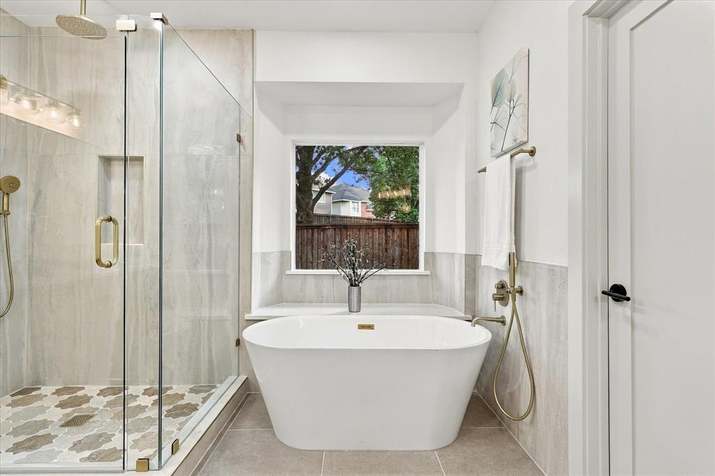 Freestanding soaking tub with brass fixtures, frameless glass shower enclosure featuring an integrated stone-tiled niche, patterned tile shower flooring, and oversized window with stone-topped ledge