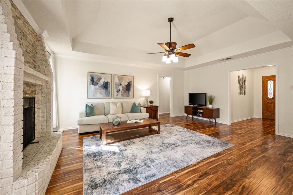 Living area featuring a floor-to-ceiling brick fireplace with stone accents, tray ceilings, wood-finish flooring, and a multi-light ceiling fan