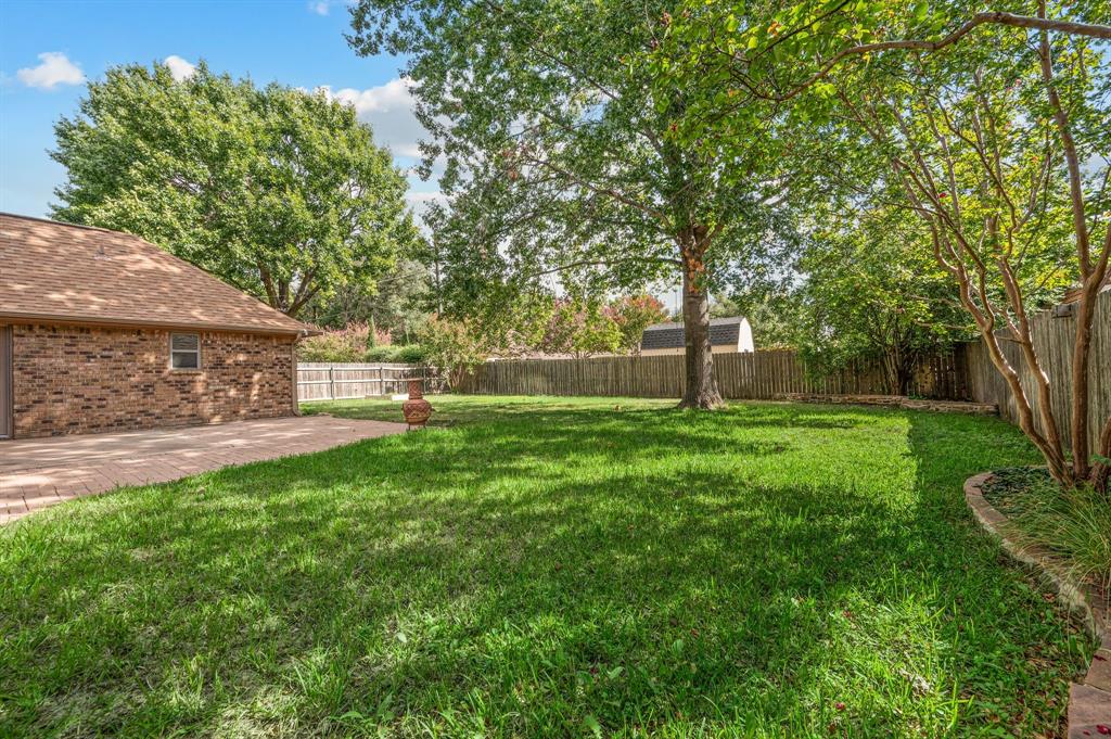 Expansive backyard featuring a brick-paved patio, manicured lawn, mature trees, and perimeter wood fencing