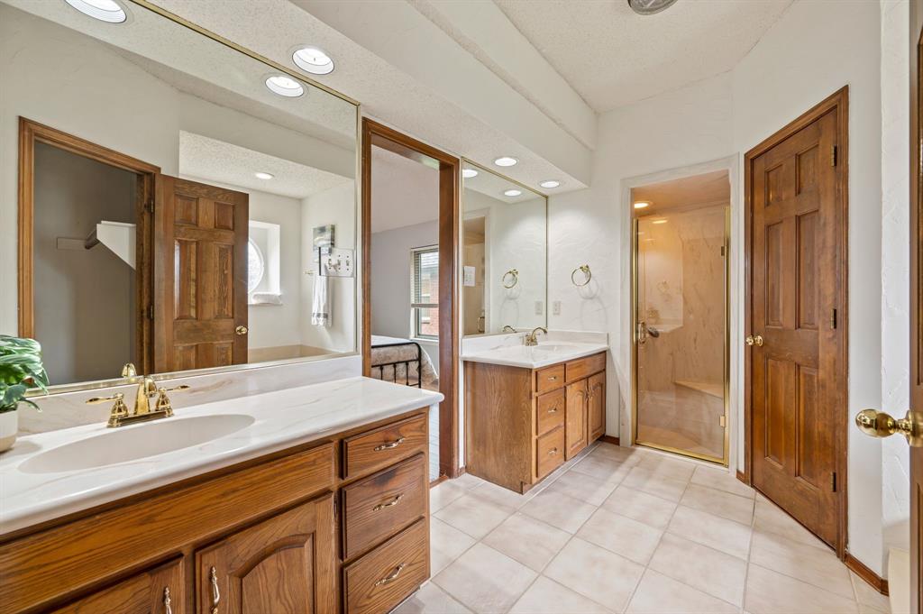 Dual-vanity cabinetry with wood finish, large wall-mounted mirrors, recessed lighting, tile flooring, and enclosed shower with glass door