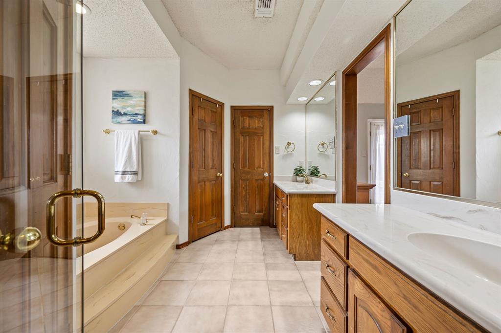 Bathroom featuring dual wood-finish cabinetry, solid-surface countertops, integrated sinks, a drop-in soaking tub, and tile flooring