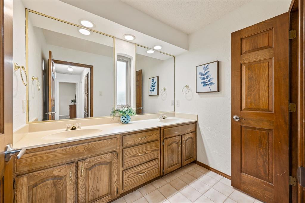 Dual-sink vanity with wood-finish cabinetry, solid-surface countertop, recessed ceiling lighting, and full-width wall mirror