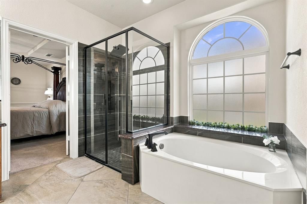 Soaking tub with stone-finish surround, glass-enclosed shower with black frame, dark horizontal wall tile, and arched window with grid detailing