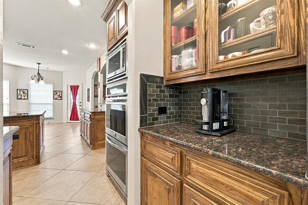 Kitchen featuring wood cabinetry with glass-front display uppers, dark stone-finish countertops, subway tile backsplash, and built-in stainless steel double ovens