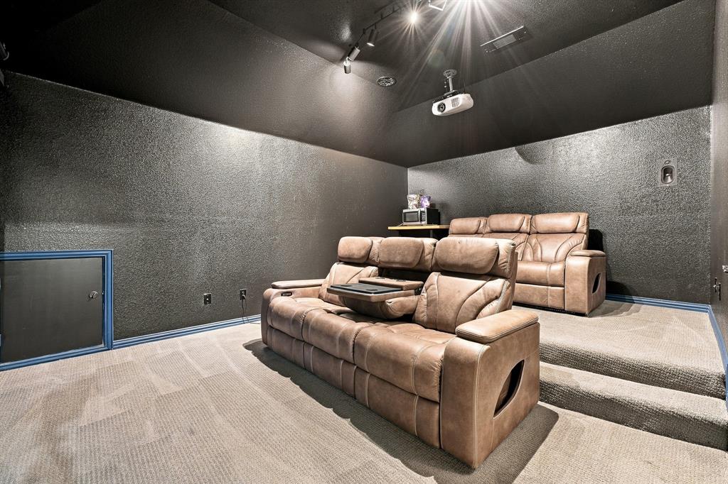 Media room featuring tiered carpeted flooring, dark textured wall surfaces, track lighting, and a ceiling-mounted projector