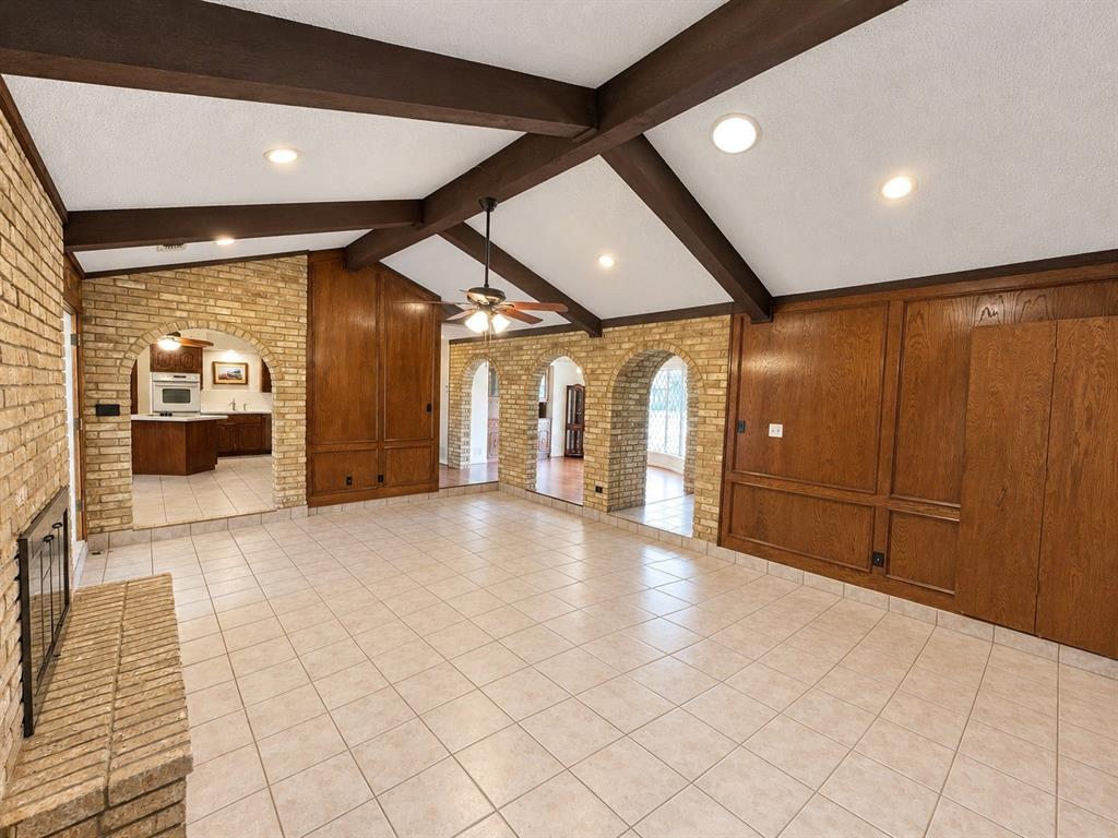 Vaulted ceiling featuring exposed dark-wood beams, brick-accented arched openings, full-wall wood paneling, tiled flooring, and a floor-to-ceiling masonry fireplace
