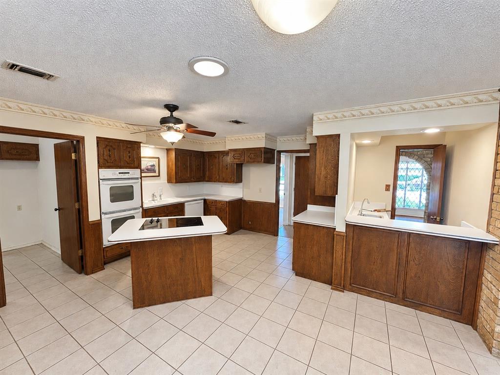 Kitchen featuring wood cabinetry, integrated double wall ovens, a center island with a cooktop, a pass-through bar, and tile flooring
