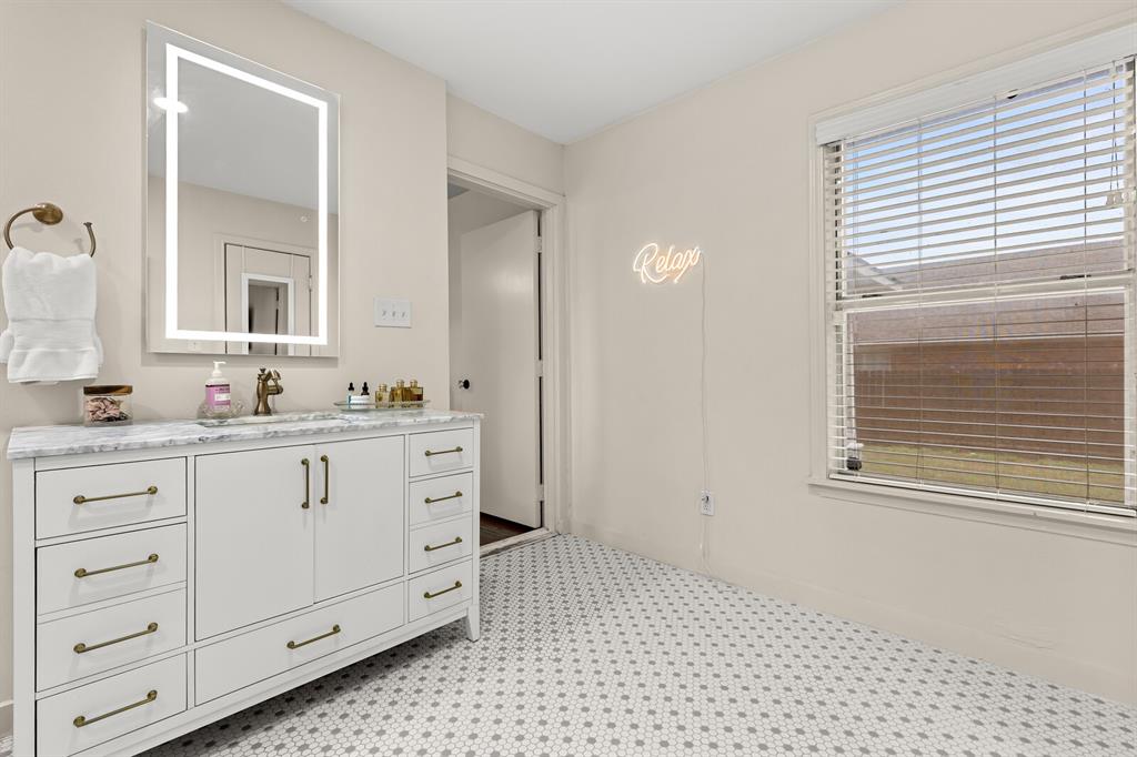 White vanity cabinet featuring brass hardware and a stone-composite countertop, illuminated by an integrated LED-frame mirror, complemented by penny-tile flooring and horizontal window blinds
