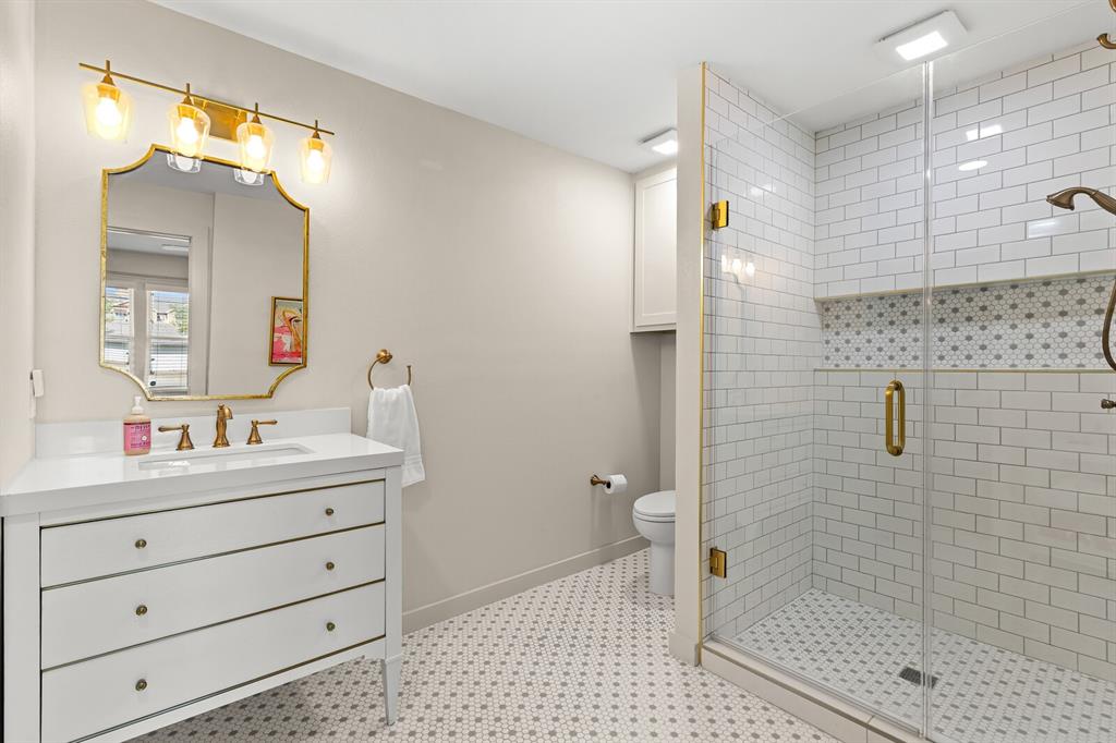 Bathroom featuring a vanity with a white countertop, gold-toned hardware, wall-mounted four-bulb light fixture, glass-enclosed shower with subway tile surround, and patterned tile flooring