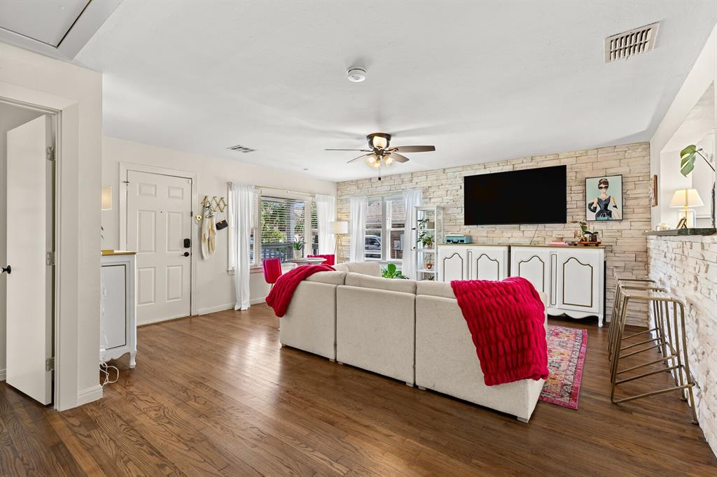 Living area featuring wood-finish flooring, a light-toned stone accent wall, multi-pane windows, and a ceiling fan with integrated light fixture