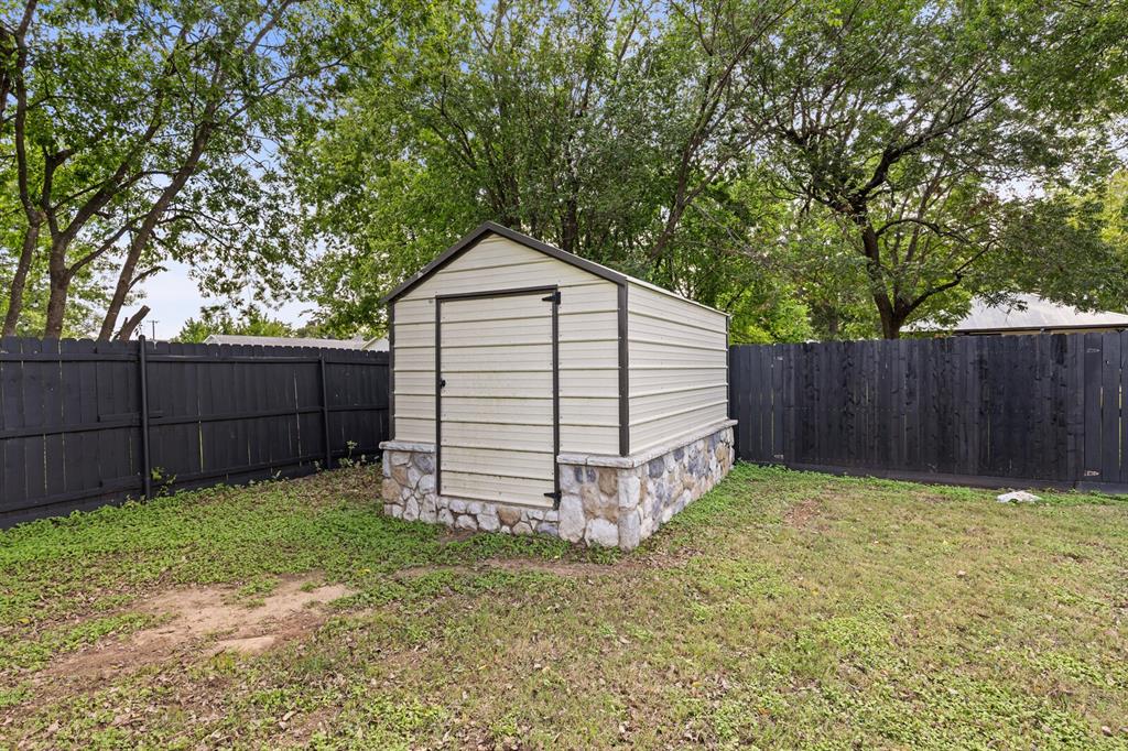 Exterior storage structure featuring a metal-sided frame, a stone masonry foundation, and a gabled roof, enclosed within a black perimeter fence