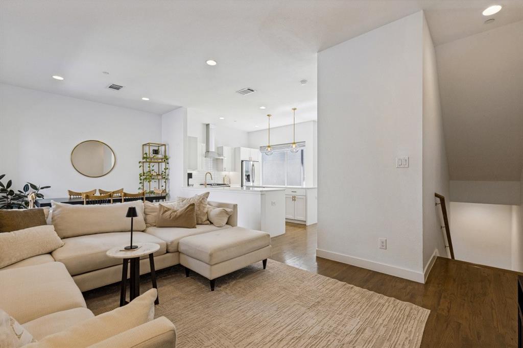 Open-concept living space featuring wood-finish flooring, recessed lighting, white cabinetry, an island with countertop seating, and a modern stairwell with a wooden handrail