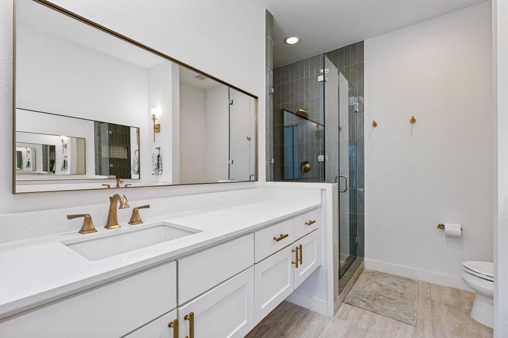 White shaker-style cabinetry with gold-tone hardware, crisp quartz countertop, expansive wall-mounted mirror, walk-in shower with vertical tile surround and glass enclosure, and wood-finish flooring
