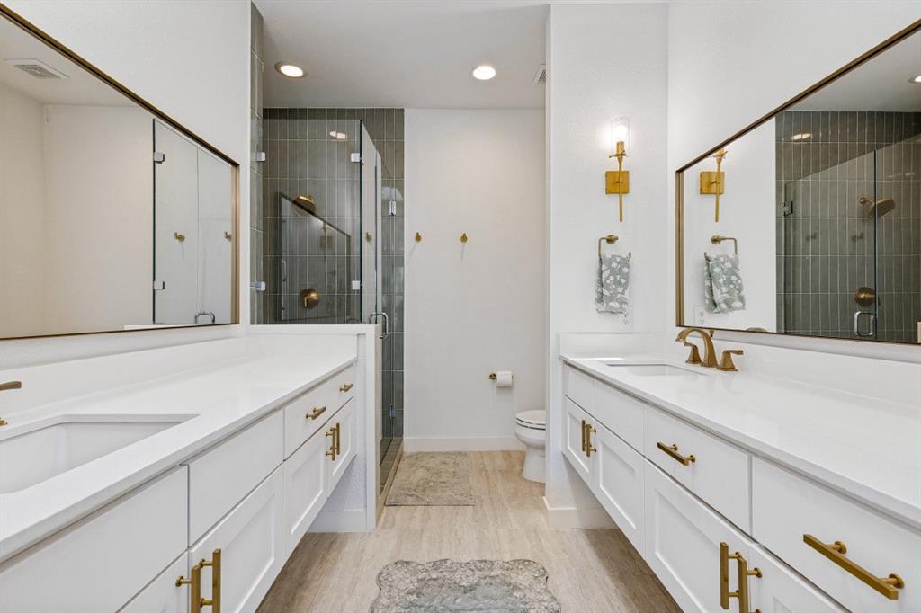 Dual vanities featuring white cabinetry, solid surface countertops, metallic hardware, large rectangular mirrors, and a glass-enclosed shower with vertical wall tiling
