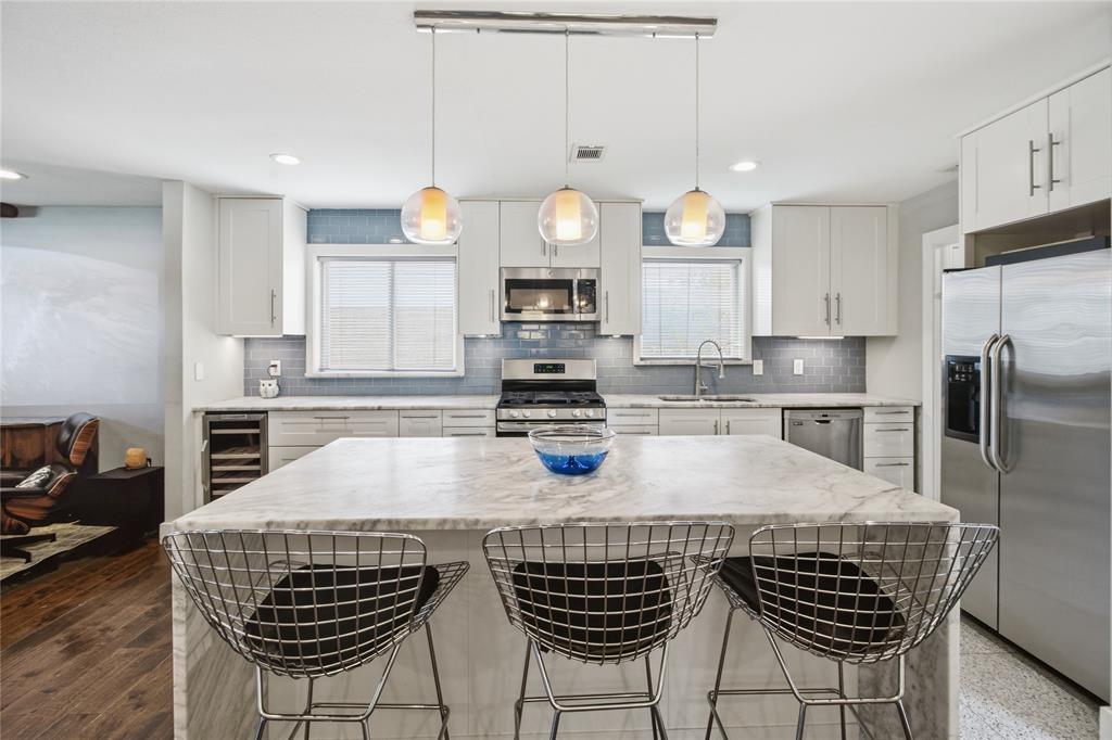 The beautifully updated kitchen features an expansive quartz waterfall island with seating for three, crisp white shaker-style cabinetry, stainless steel appliances, and a soft blue subway tile backsplash. Three glass pendant lights complete the contemporary design.