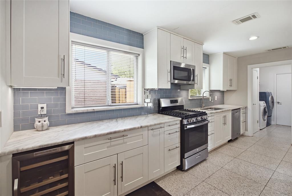 The thoughtfully designed kitchen features white shaker-style cabinetry, quartz countertops, a glossy blue subway tile backsplash, and stainless steel appliances. A gas range, built-in beverage cooler, generous counter space, and natural light from two windows combine style with everyday functionality.