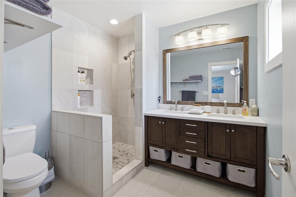 The primary ensuite bathroom features a spacious walk-in shower with tile surround, pebble-style flooring, and recessed storage niches. A double-sink vanity with dark wood cabinetry, generous counter space, and open shelving provides everyday convenience in a contemporary setting.