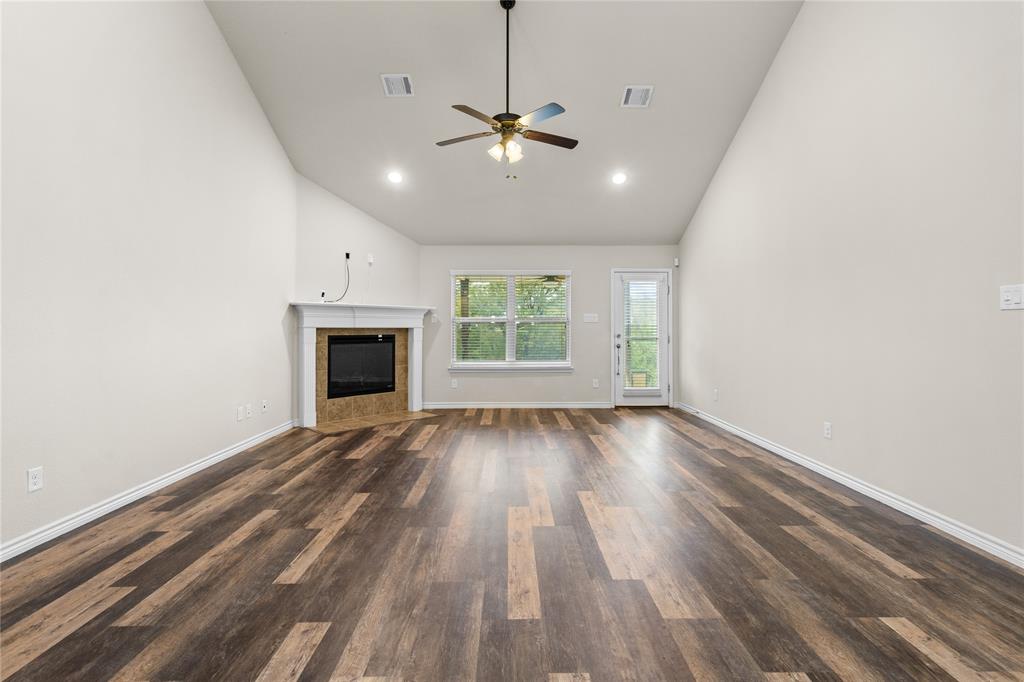 Vaulted ceiling living area featuring wood-finish flooring, a fireplace with a white mantel, a ceiling fan with integrated lighting, and glass-paneled exterior door