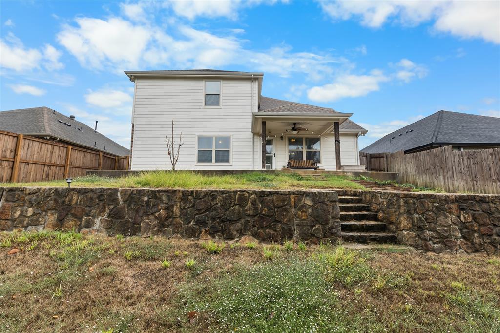 Rear exterior featuring a two-story light-colored siding facade, a covered patio with dark support pillars, and a stone retaining wall with built-in stone steps