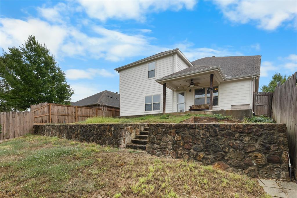 Two-story light-colored siding exterior featuring a covered patio, tiered natural stone retaining wall, integrated stone staircase, and wood privacy fencing