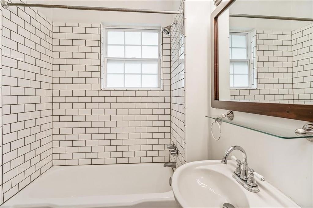 Bathroom featuring white subway tile surround, integrated bathtub, grid-pattern window, pedestal sink with chrome faucet, and wood-framed mirror
