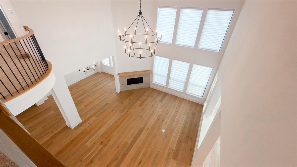 Double-height ceiling, wood-finish flooring, stone-surround fireplace with a wood mantel, multi-tier metal chandelier, and curved balcony with a metal railing