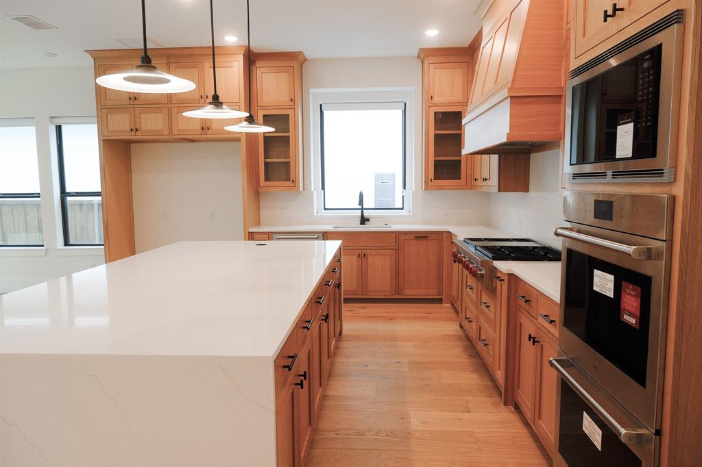 Kitchen featuring custom light wood cabinetry, white stone-surface countertops, a large center island, stainless steel wall-mounted oven and microwave, and wood-finish flooring