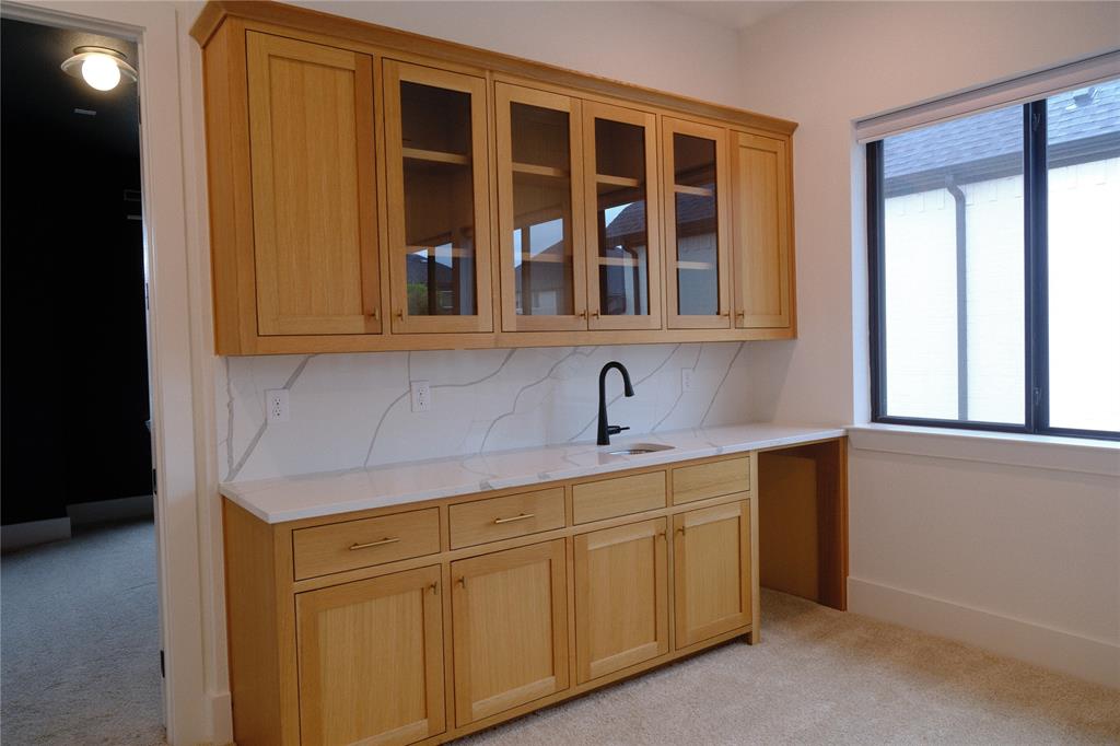Custom cabinetry featuring glass-front upper doors, stone-finish countertops, full-height decorative backsplash, integrated sink with matte black faucet, and built-in workspace