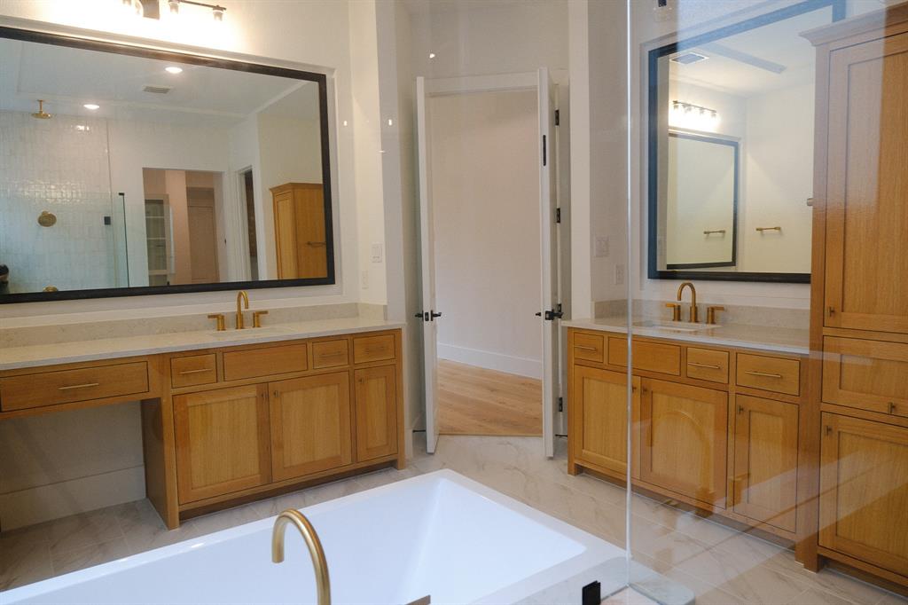 Dual wood-finish vanities with light stone countertops, integrated cabinetry, expansive framed mirrors, and a white freestanding tub with gold-tone fixtures