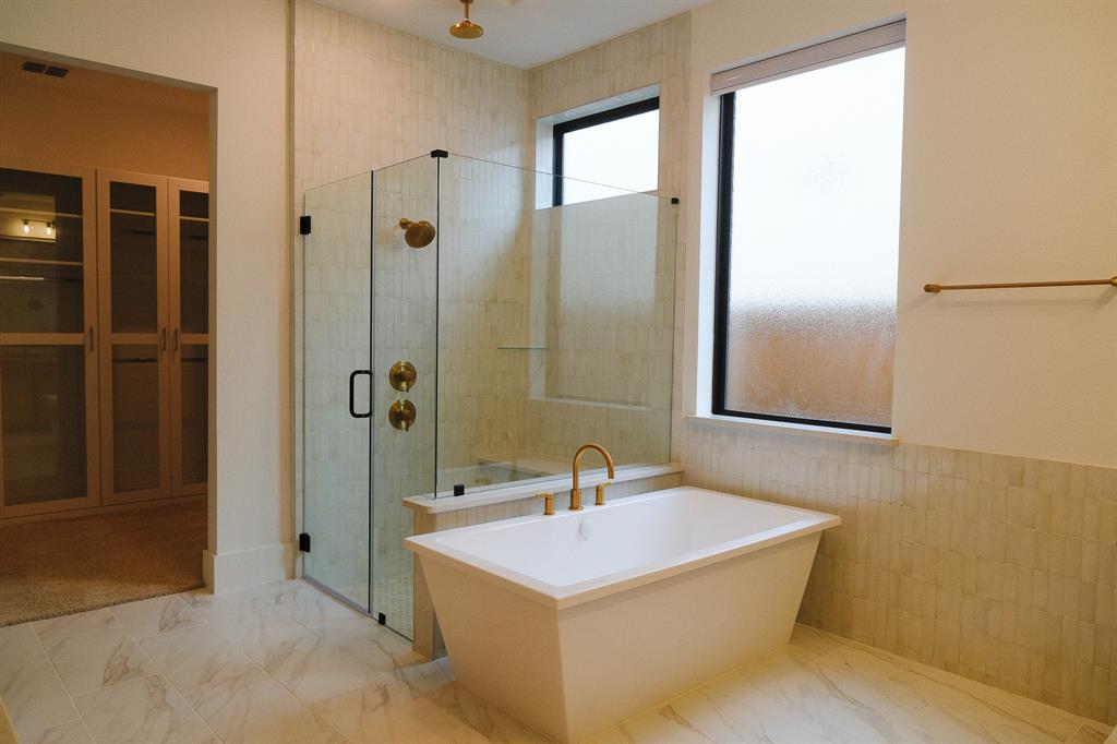 Freestanding soaking tub featuring gold-tone hardware, glass-enclosed shower with vertical wall tile, light-toned stone-patterned tile flooring, and integrated built-in cabinetry