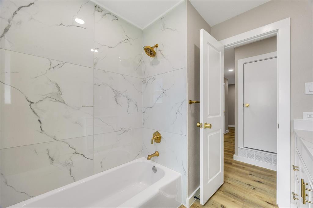 Full-height wall tile with veined pattern, brass-finish fixtures, white soaking tub, wood-finish flooring, and shaker-style cabinetry